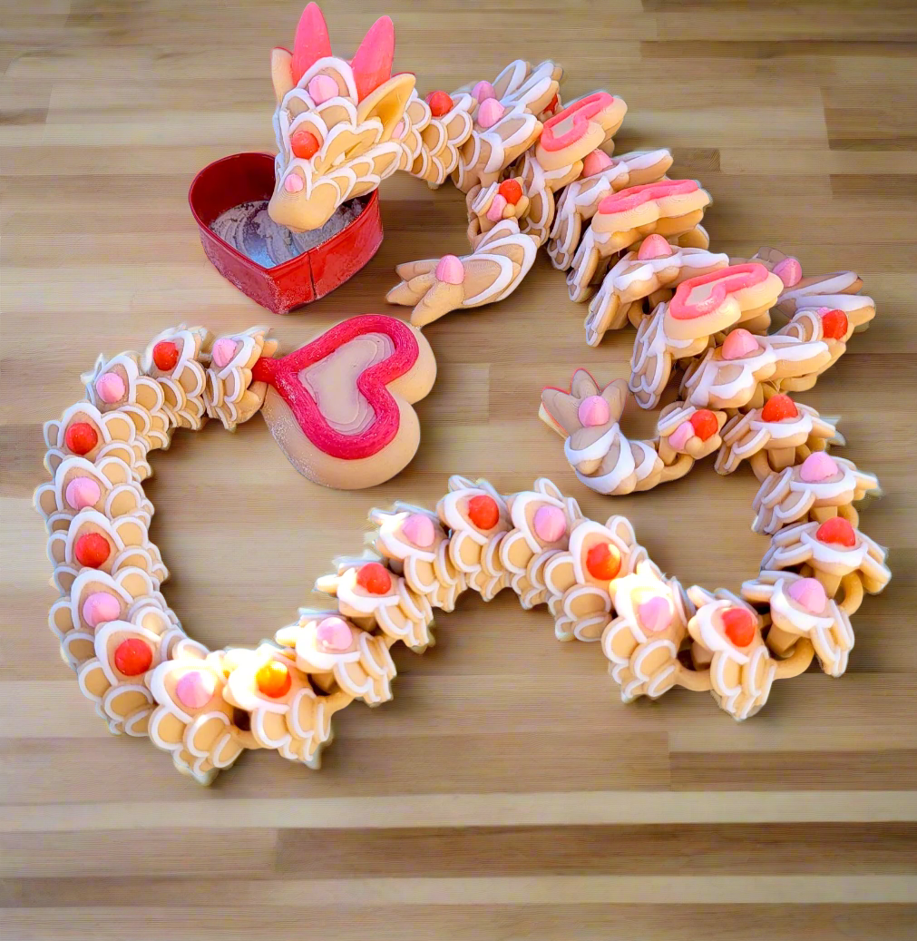 A 3D printed, articulated flexi dragon cookie toy with heart-shaped wings and frosting-like details, positioned on a surface with a background story text that reads 'Heart Cookie Dragon'.
